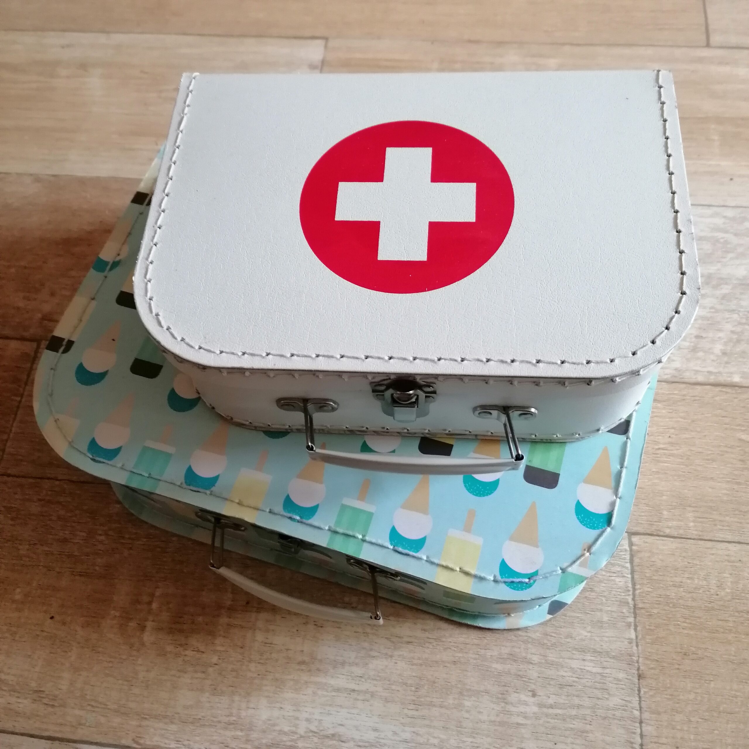 Twee kinderkoffers op elkaar. De onderste is lichtblauw met ijsjes erop, gele waterijsjes en blauw/witte softijsjes met een hoorntje. De bovenste is een witte met een rode cirkel erop, waar een wit kruis in staat.